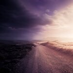 Ein Feldweg führt durch eine Landschaft, die links in dunklen Wolken und rechts in hellem Sonnenlicht liegt